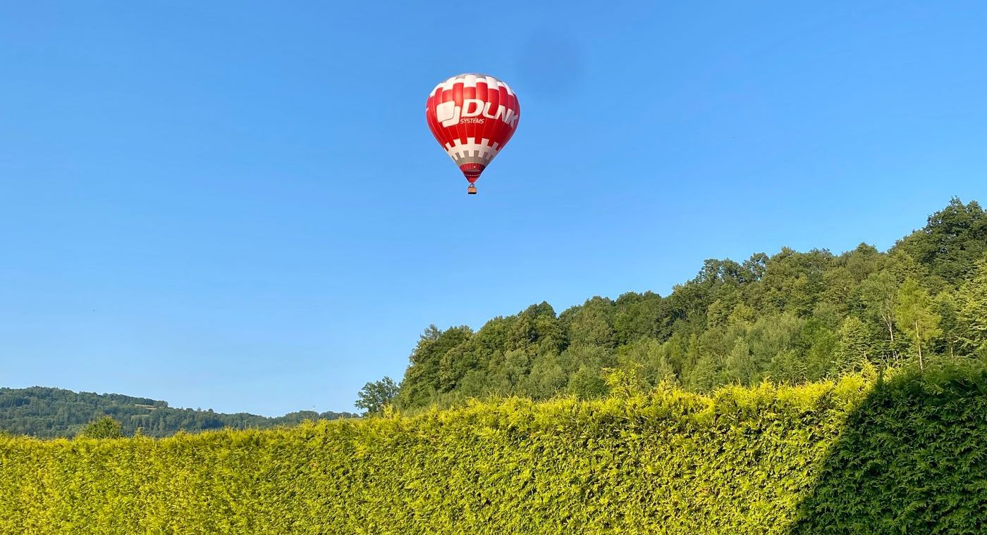 Une montgolfière seule dans un ciel dégagé, au-dessus d’une haie et de collines boisées