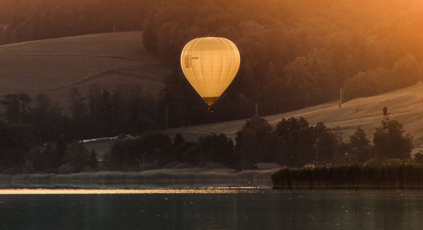 Une montgolfière et son reflet sur un lac, à contre-jour, entre collines boisées
