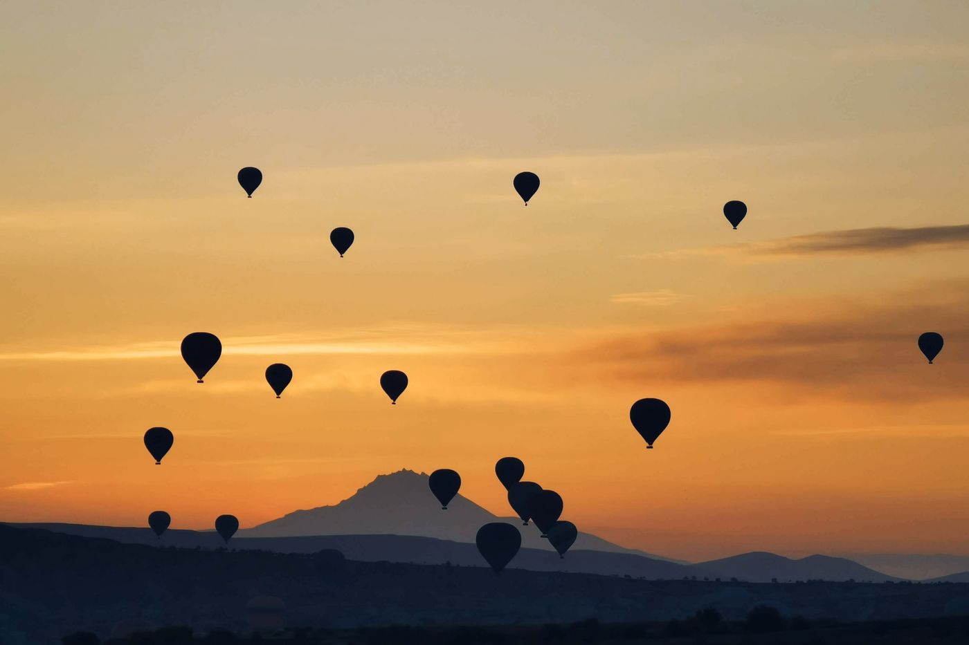 Une multitude de montgolfières en silhouette dans un ciel orangé au coucher du soleil, au-dessus d’un relief montagneux